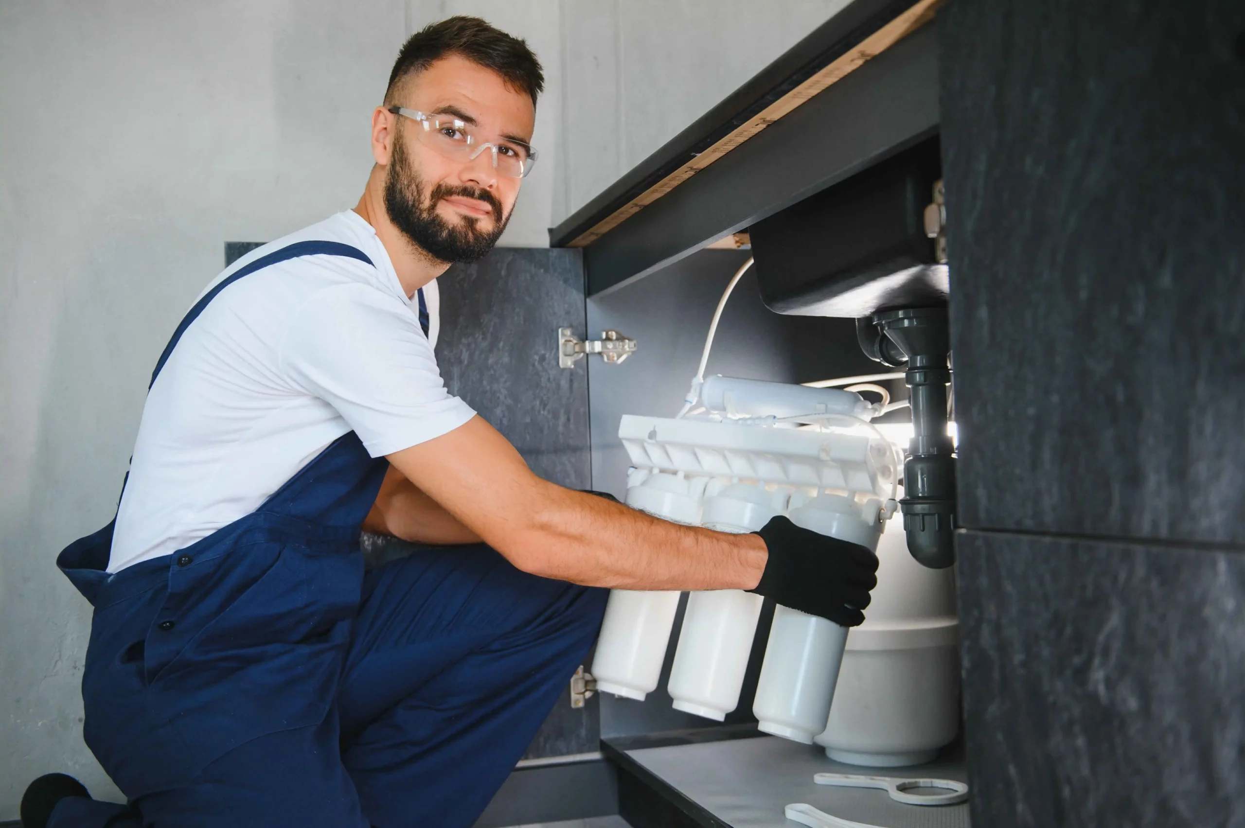 A male technician in overalls and safety glasses kneeling under a sink, installing a water filtration system.
